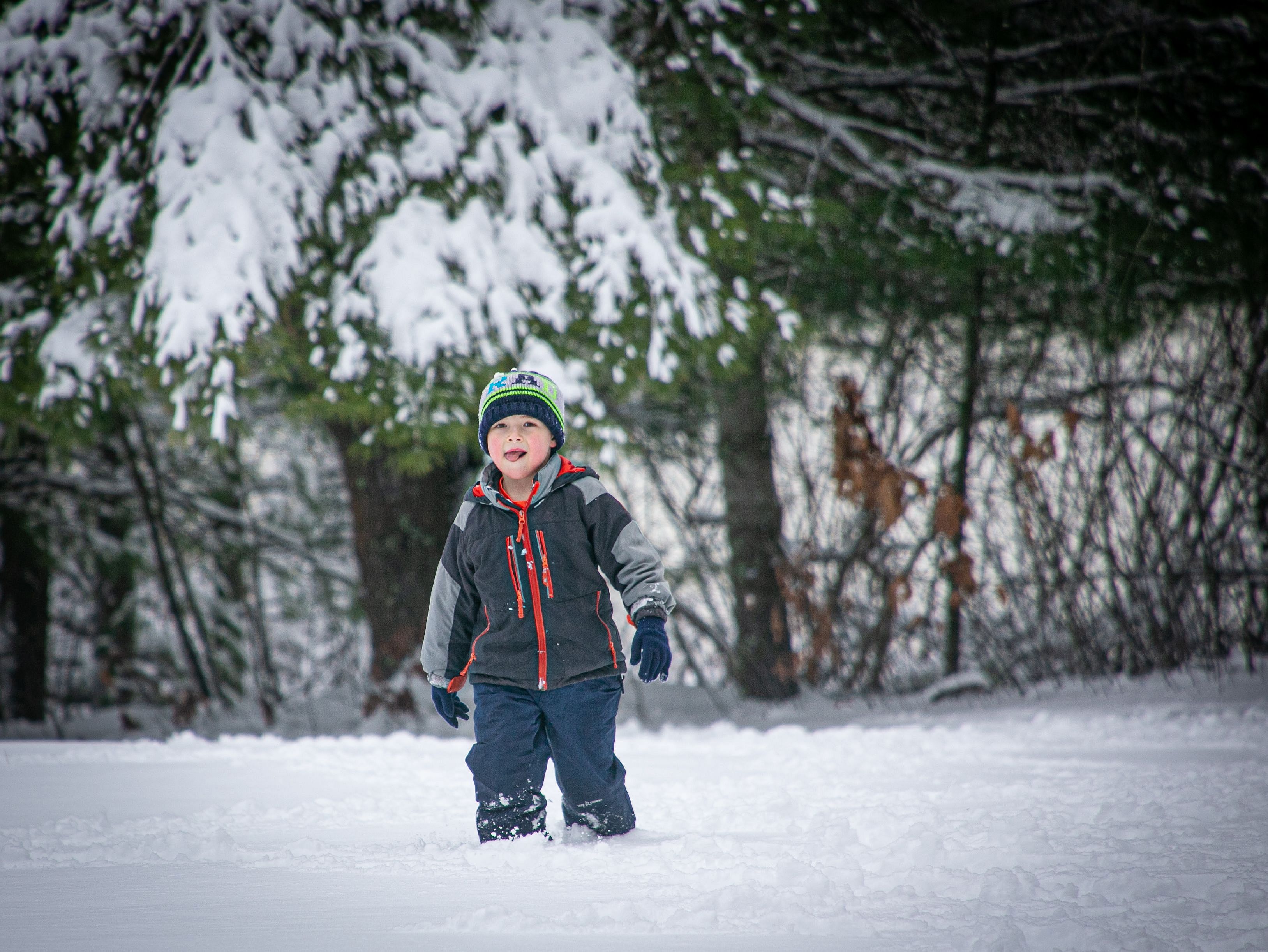 A little kid standing in front of pine trees and wearing a snow suit, licks a snowflake off his chin.