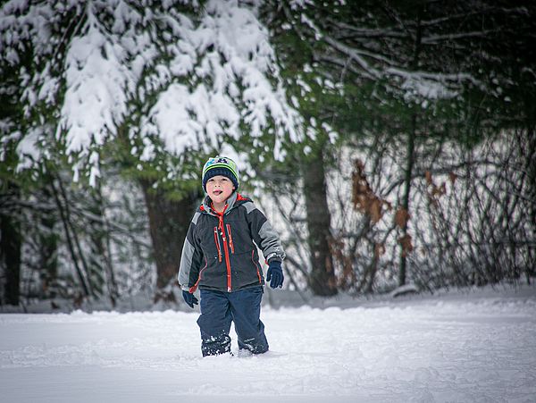 A little kid standing in front of pine trees and wearing a snow suit, licks a snowflake off his chin.