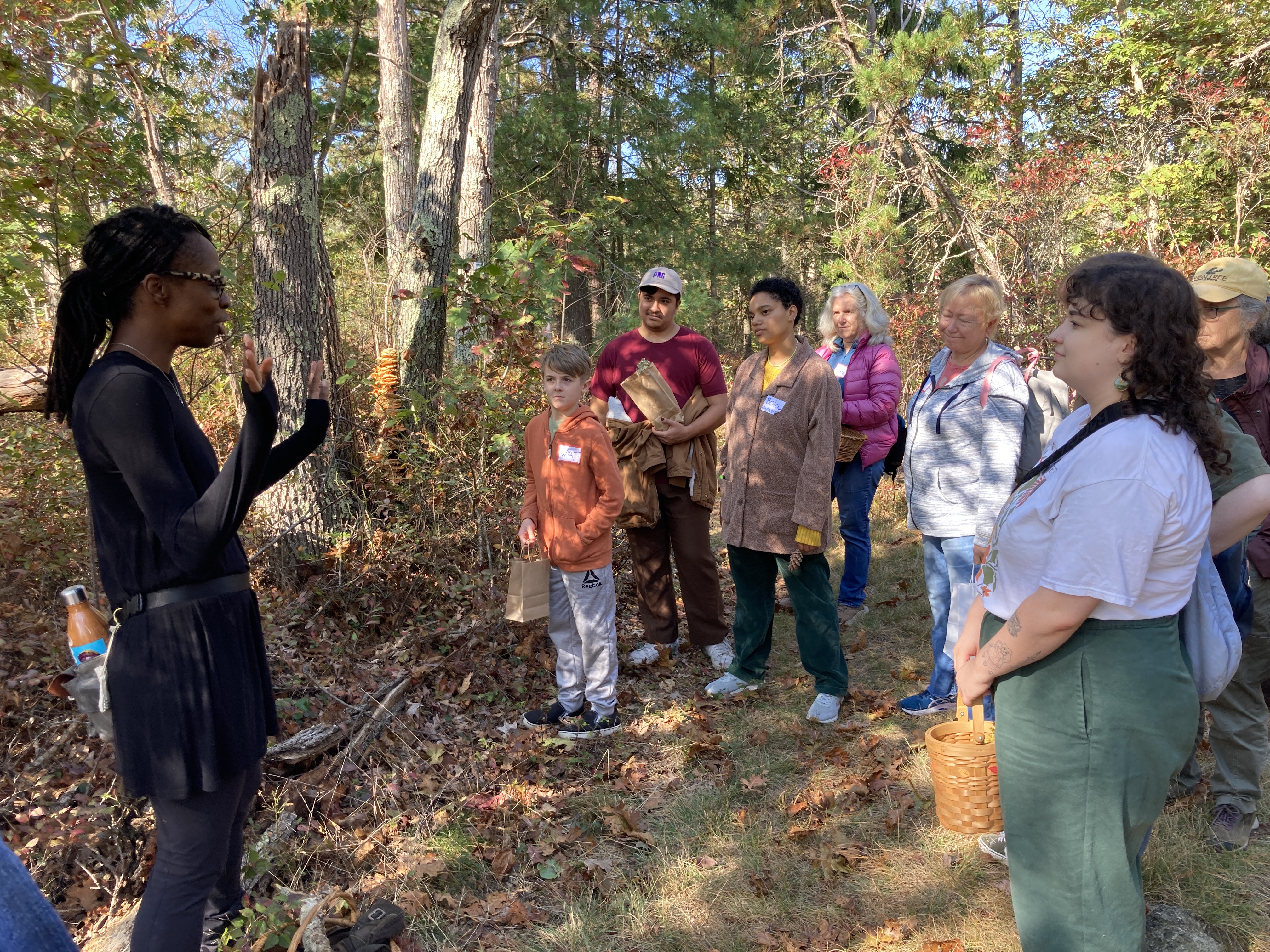 A photo of Maria Pinto, the mushroom expert, explaining identifying characteristics of mushrooms to a group of interested people on a wooded trail.